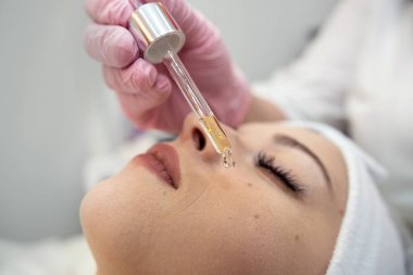 Kozmetik uzmanı, spa merkezindeki genç güzel kadınının yüzüne pipetle birkaç damla nemlendirici yağ sürüyor. Yüz bakımı prosedürü