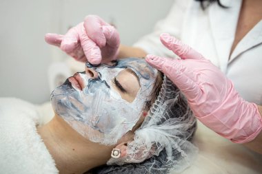 Kozmetik uzmanı, kaplıca merkezindeki kadın müşterisini gençleştirici maskeyle yüz masajı yapıyor. Yüz dermatolojisi maskesi. Profesyonel cilt bakımı