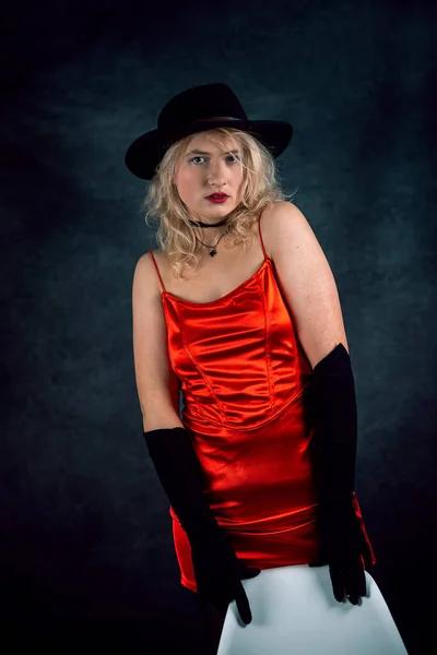 pretty young blond woman wear red evening dress and black hat isolated on dark background. lifestyle
