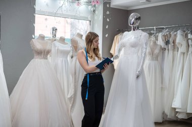 Pretty  female dressmaker with diary and pen posing near bridal wear in wedding fashion boutique