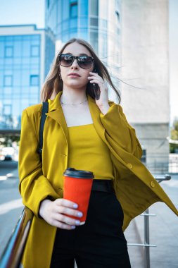 A business woman with a cup of coffee is talking on her smartphone while standing in the middle of the city center. The concept of a woman who earns money