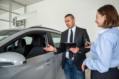  Galeride modern bir arabanın yanında duran genç ve güzel bir bayan satıcıdan yardım alarak en iyi otomobili seçiyor. satın alma kavramı
