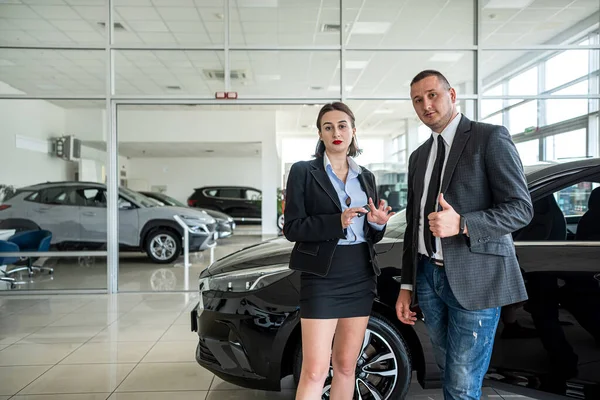 salesperson in a car dealership and a buyer show thumbs up as a sign of satisfaction with the purchase of a car. buying a car at a car dealership. the concept of buying and selling cars.