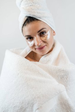 Havlu giymiş genç beyaz bir kadın beyaza izole edilmiş yüzüne hidrojel bantlar uyguluyor. Cilt bakımı kozmetik prosedürleri