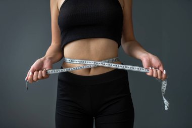 Gri arka planda izole edilmiş bant ölçüsüyle ideal belini ölçen gülümseyen güzel bir kadın. Arka planda stüdyoda bir kadın var. Diyet Sağlıklı Yaşam Tarzı