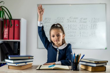 Bir ilkokul öğrencisi sınıfta öğretmenin sorusuna cevap vermeye hazır olduğu için elini kaldırır. Akıllı küçük bir kız sınıftaki bir masada oturuyor. Eğitim kavramı.