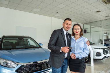 Beautiful man and woman couple holding key to their new car looking at camera and smiling in car showroom. purchase of a family car. new car showroom