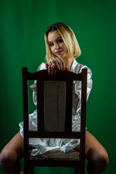 白衣を着た魅力的な若いスリムなセクシーな女性は プレーンな背景と笑顔で木製の椅子の上にスタジオに座っています 隔離されたスタジオ 肖像画 — ストック写真