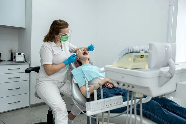 ヨーロッパの若い女性は医者の椅子に座っていますが 女性の歯科医は歯科クリニックで歯を固定しています 歯医者の健康な歯 — ストック写真
