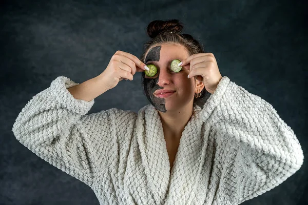 Jong Meisje Witte Badjas Zwarte Klei Masker Houden Komkommer Plakjes — Stockfoto
