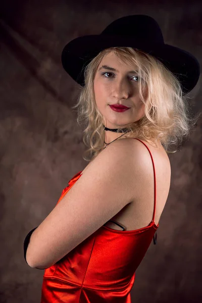 暗い背景に隔離されたスタジオで赤エレガントな夜のドレスと黒の帽子のポジションで美しいブロンドの女性 ファッションルックスタイルのコンセプト — ストック写真