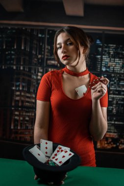 Poker masasının yanında kırmızı elbiseli güzel bir kız elinde bir şapka ve renkli cipsler tutuyor. Poker konsepti Kadın