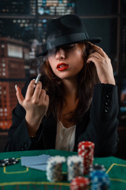 young girl stylishly dressed smokes a cigarette, blows smoke and plays cards on a table on a green cloth. casino. poker