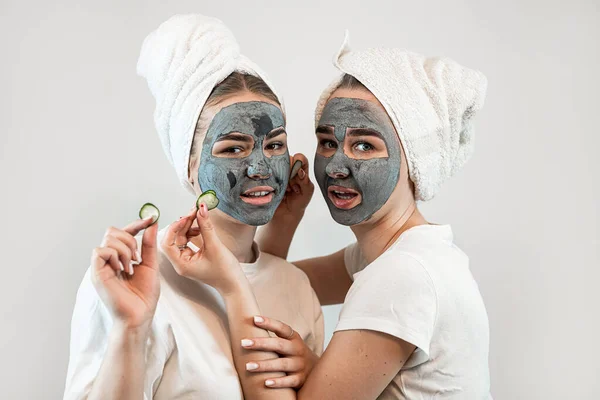 黒い粘土の顔のマスクを作り 白に隔離された手にキュウリのスライスを保持する2人の若い女性 友情の概念 スキンケアと治療のための自然な化粧品顔マスク — ストック写真