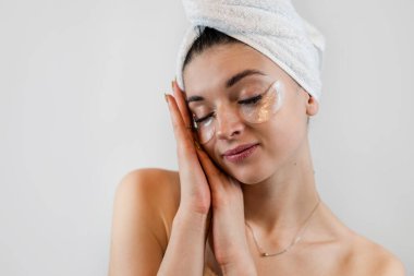 Havlu giymiş genç bir kadın beyaza izole edilmiş gözlerinin altında kolajen hidrojel yama uyguluyor. Kırışıklık önleyici göz bantları. Cilt bakımı kavramı.