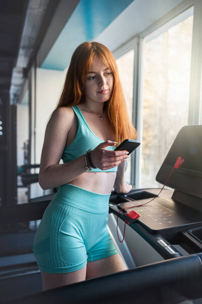 Young red-hair woman stand near treadmill trainers using mobile cell at gym. Workout, lifestyle