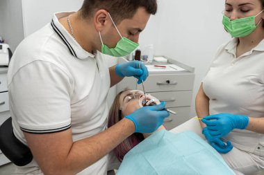 Mavi eldivenli dişçi ve bayan asistan dişçi aletleriyle çalışıyor. Dişçinin ofisinde dişlerini tedavi eden güzel bir genç kadın. Dişçi koltuğundaki hasta..