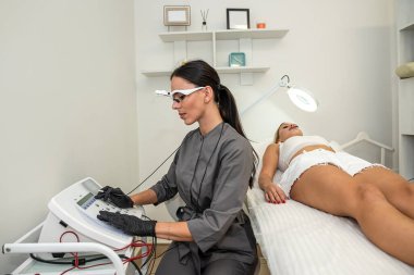 Kozmetik uzmanı elektroepilasyon işlemi için her şeyi hazırlar, genç bayan müşteri yatağa uzanıp bekler. Güzellik ve temiz ten kavramı