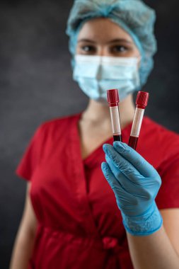Kırmızı üniformalı, koruyucu eldivenler giyen, maske takan ve kırmızı kanla test tüpü tutan genç bir hemşire. Sağlık laboratuvarı kavramı