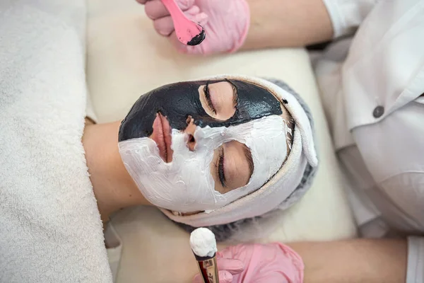 Stock image The cosmetologist applies a black and white mask on half of the face at spa salon. Skin care and treatment. Anti-wrinkle cosmetic procedure 