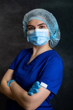 Koruyucu tıbbi maskeli genç kadın doktor portresi koyu arka planda mavi üniforma giyer. Sağlık Hizmetleri
