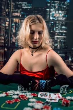 Kırmızı gece elbisesi giymiş duygusal genç bir bayan kumarhanede yeşil bir bezin üzerinde kağıt oynuyor. Poker oynayan bir kadın. kırmızı elbise