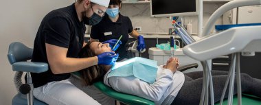 Erkek dişçi, dişçi kliniğinde gülümseyen genç bir kadını ameliyat eder ve kadın asistan ona yardım eder. Oral bakım konsepti. Sağlıklı dişler fırçalıyor.