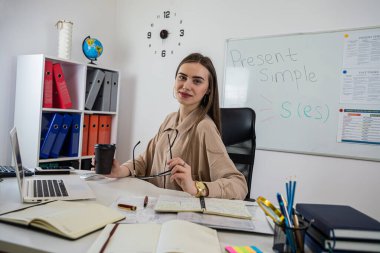 Genç bir öğretmen tüm kuralların yazılı olduğu tahtaya yakın masada otururken İngilizce dilbilgisi kurallarını söyler..