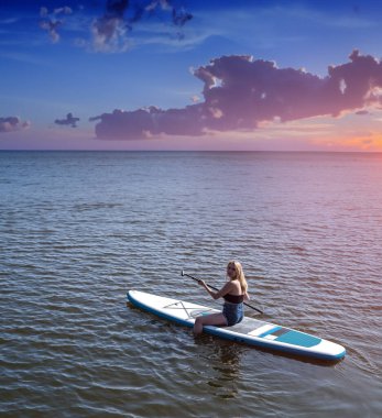 Genç, sıska bir kadın, yaz mevsiminde SUP kürekleri üzerinde ayakta duruyor. Sağlıklı yaşam tarzı