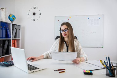 Gülümseyen kadın öğretmen çalışma masasında dizüstü bilgisayarla oturuyor ve yazı tahtası yakınındaki dil bilgisi kurallarını anlatıyor.