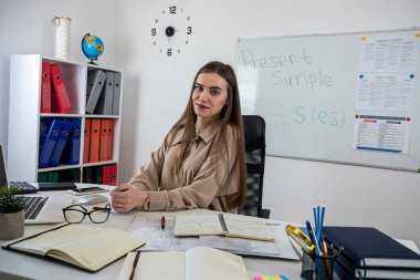 Gülümseyen kadın öğretmen çalışma masasında dizüstü bilgisayarla oturuyor ve yazı tahtası yakınındaki dil bilgisi kurallarını anlatıyor.