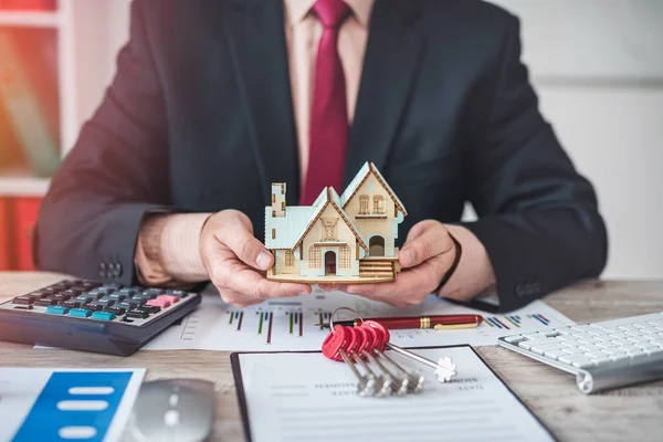 house salesman man in jacket sitting with documents for house sale contract and keys to house model. real estate trading concepts. property insurance