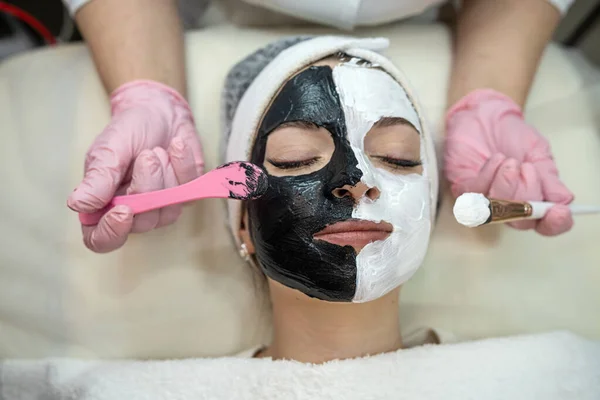 young woman with black and white clay cosmetic mask on half face lying a bad on spa salon. Skin care and treatment, lifestyle