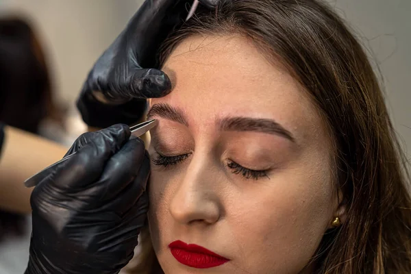 lashmaker makes a beautiful eyebrow contour with tweezers. female face close-up. A beautician does an eyebrow procedure in a beauty salon for a girl. Beauty industry concept.