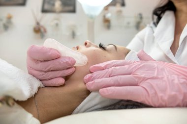 Kozmetik uzmanı müşterinin yüzüne beyaz guaj kazıyıcı sürerek tıbbi işlem yapıyor. Sağlık yaşam tarzı