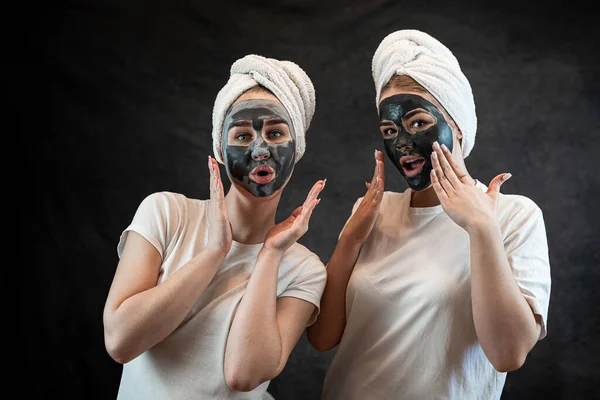 Two happy friends wear white tshirt towel applying black clay facial mask isolated on black. Skin care for all ages. SPA and wellness.