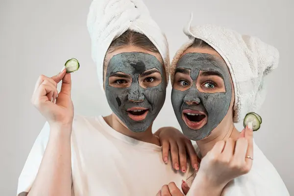 Two girlfriend or sister wear white towel with black clay mask hold pieces of cucumber isolated on white. Skin care and treatment