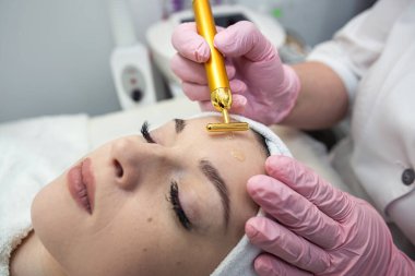 Kozmetik uzmanının masasında uzanan ve yüzünde titreşim masajıyla prosedür uygulayan genç bir kadın. Kaplıcada cilt bakımı