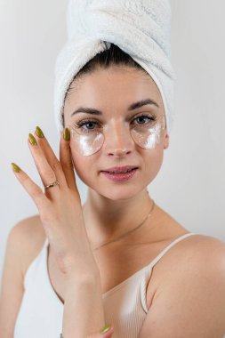 Beyaz tenli bir kadın, beyaza izole edilmiş gözlerinin altında hidrojel bantlarla parlayan cilt yüzeyine dokunuyor. Kırışıklık önleyici göz bantları