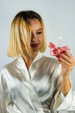 Pijamalı güzel genç bir kadın bir kekin üzerine doğum günü mumu üflüyor. Tipik bir stüdyo arka planında izole edilmiş. Doğum günün kutlu olsun