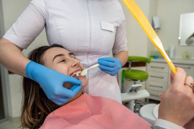 Kadın dişçi elinde renk örnekleri tutuyor ve hastasının aynaya bakması için bir gölge seçiyor. Diş bakımı ve hijyen