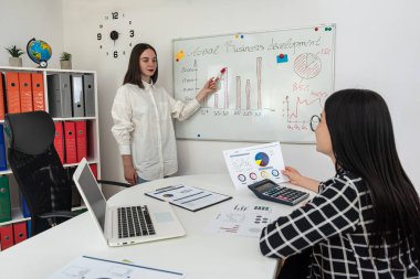 Çizelgenin yanında duran iki genç kadın ofiste iş grafiğini tartışıyor. Takım çalışması.