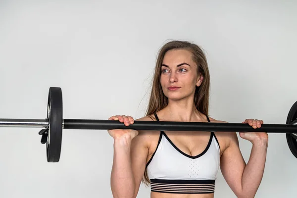 Intense training in the gym a stressed young female athlete is ready to perform an exercise with a heavy barbell on a white plain wall. isolated gaining muscle mass