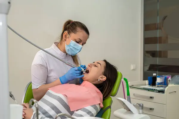 Professional female dentist using sterile dental drill and mouth mirror treating female patient teeth at dental clinic. Health care