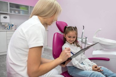 Küçük kız, dişçide çocuğun şikayetlerini kaydeden bir kadınla konsültasyonda oturuyor. Pediatrik diş hekimliği okuyan dişçi çocuk
