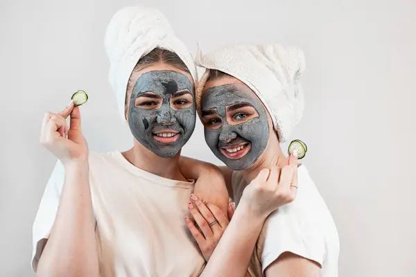 Two young woman making black clay facial mask and holding cucumber slices on hand isolated on white. Friendship concept. Natural cosmetic facial mask for skin care and treatment