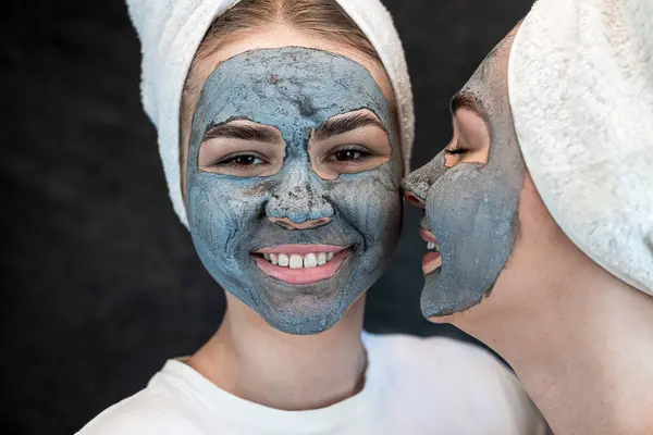 Two happy friends wear white tshirt towel applying black clay facial mask isolated on black. Skin care for all ages. SPA and wellness.