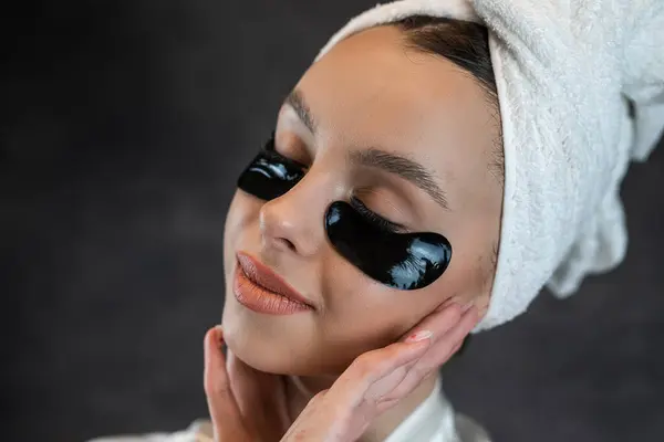 Beauty woman in towel applies black collagen patches under eyes isolated on black.  Skincare, anti fatigue eye mask