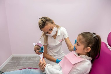 female dentist shows the treated teeth to the little girl patient in front of the mirror. children's dentistry. beautiful little smile. mirror in hands