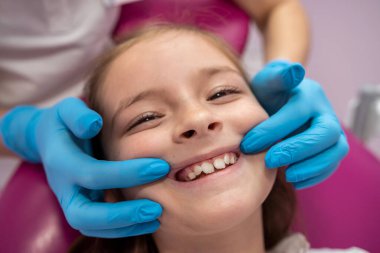 Doktor küçük bir kızın ağız boşluğunu inceliyor. Diş çürüklerini kontrol etmek için ağız aynası kullanıyor. Dişçi kadın gülümseyen tatlı bir kız için muayene prosedürü uyguluyor.